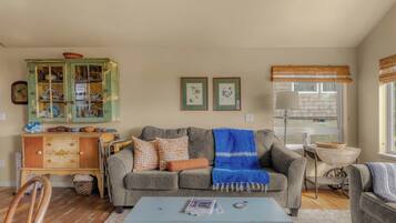 A living room with a grey sofa, a blue blanket, a wooden cabinet with glass doors, and a window with a bamboo blind.