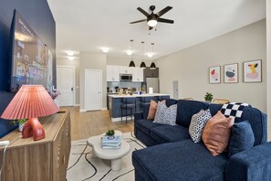 A modern living room with a sofa, a coffee table, and a TV. There is a kitchen in the background with a bar counter. - Luxe 2BR Downtown Fort Worth; Balcony, Sleeps 6 ; Near Sundance Sq & Dickies (Fort Worth)
