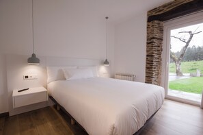 A bedroom with a large bed, two bedside tables, a nightstand, a radiator, and a window with a view of a tree and grass. - TWO-BEDROOM APARTMENT (castroverde)