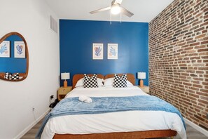 Ein Hotelzimmer mit blauer Akzentwand, einem Holz-Kopfteil, karierten Kissen, einem Bett mit blauer und weißer Decke, einem weißen Handtuch, einem Nachttisch aus Holz, einer Lampe und einem Deckenventilator.