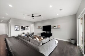 Ein modernes Wohnzimmer mit einem grauen Ecksofa, einem Flachbildfernseher auf einem schwarzen Standfuß und einem Deckenventilator.