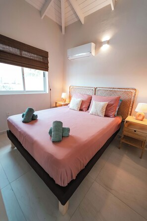 A bedroom with a bed, two bedside tables, a window with blinds, an air conditioning unit, and a lamp. - Casa Sinku Solo - Bohemian Bungalow Curacao (Willemstad)
