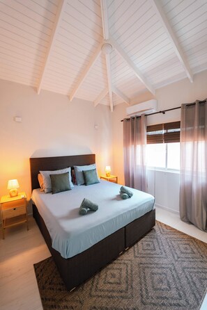 Ein Schlafzimmer mit einem großen Bett, zwei Nachttischen, einem Fenster mit Vorhängen und einer Decke mit freiliegenden Balken.