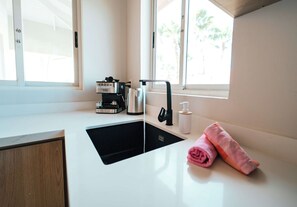 A modern kitchen with a black sink, a coffee machine, and a window with a view of palm trees. - Casa Sinku Solo - Bohemian Bungalow Curacao (Willemstad)