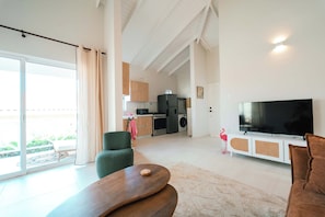 Ein modernes Wohnzimmer mit einem Flachbildfernseher, einem grünen Sessel, einem Holztisch und einer Küche im Hintergrund.