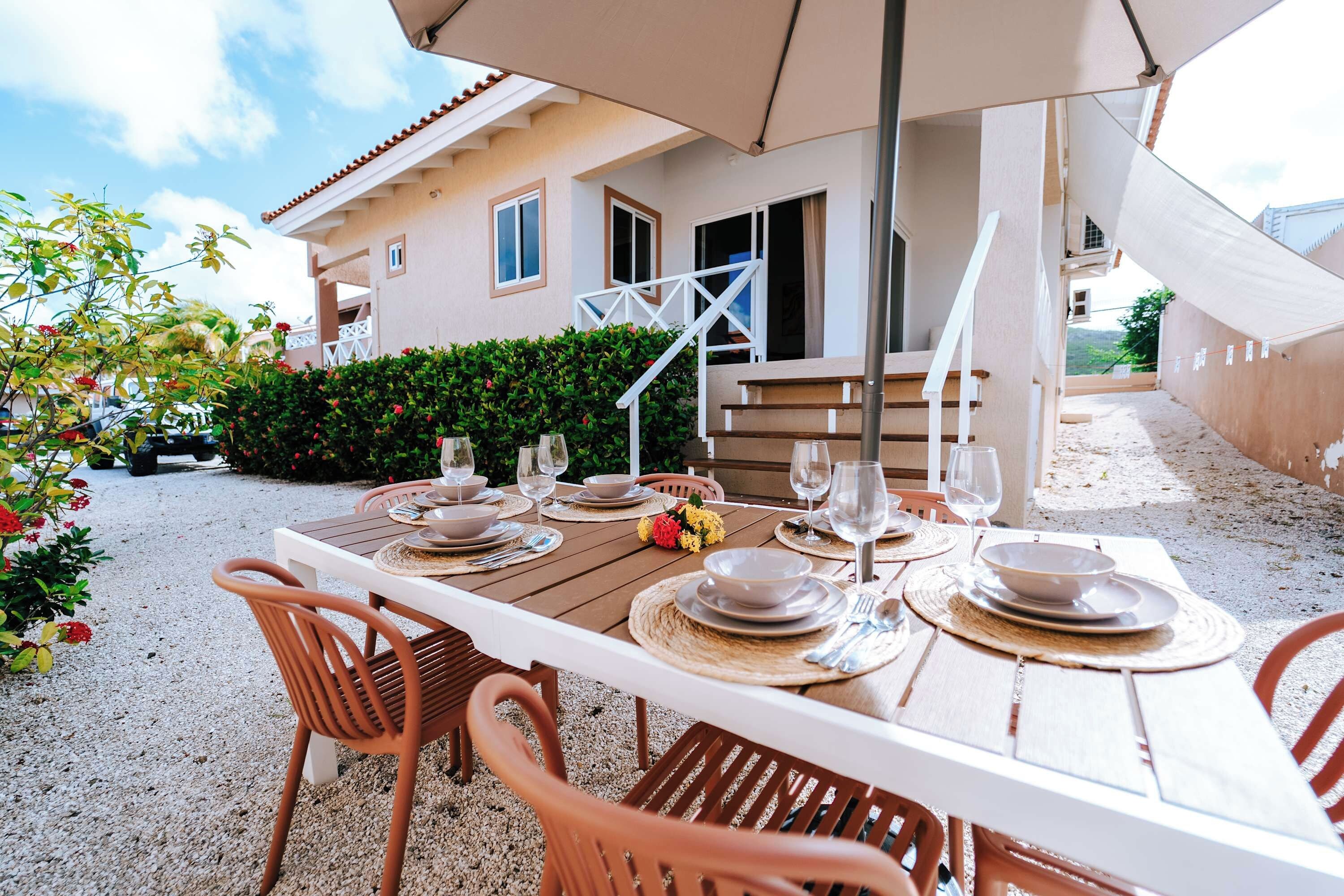 Un espace de restauration en plein air avec une table dressée pour quatre personnes, entourée de chaises, et un parasol offrant de l’ombre.