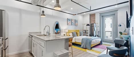 A modern apartment with a kitchen, a central island, a bed with a checkered headboard, and a large window.