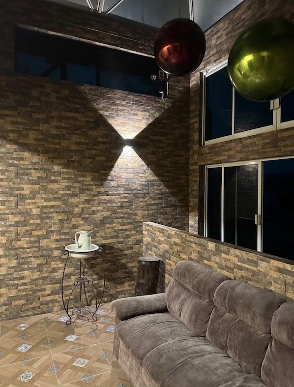 A room with a brick wall, a window, a sofa, a small table with a pitcher, and a hanging light fixture.