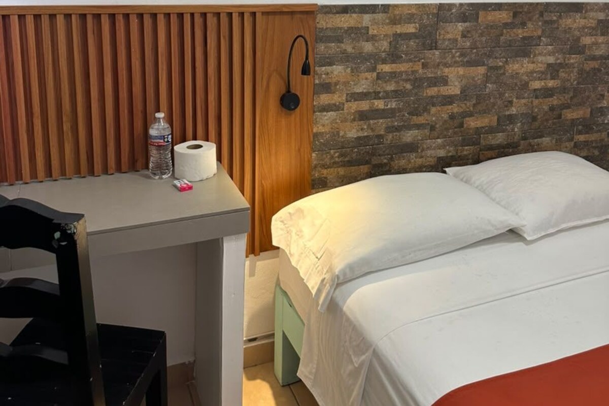 A hotel room with a bed, a desk, a chair, a wooden slat wall, a brick-patterned headboard, and a wall-mounted light fixture.