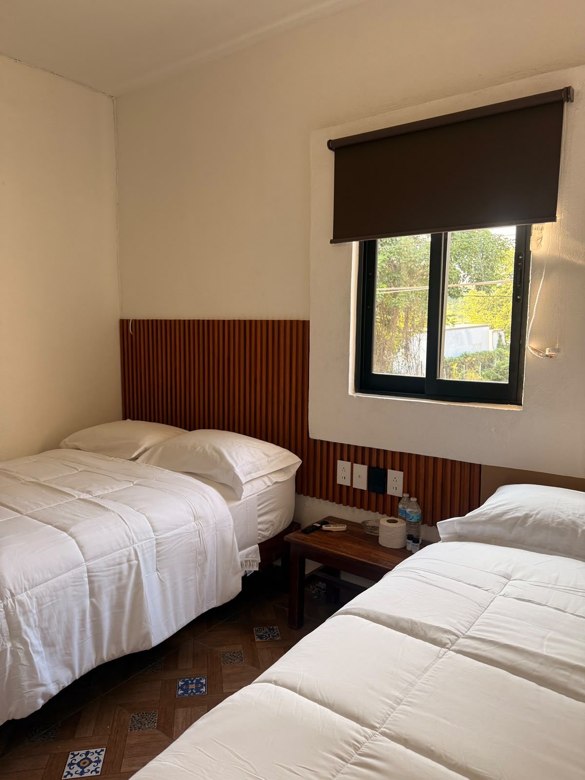 A hotel room with two single beds, a wooden headboard, a window with a blind, a bedside table with a remote control, and a wall socket.