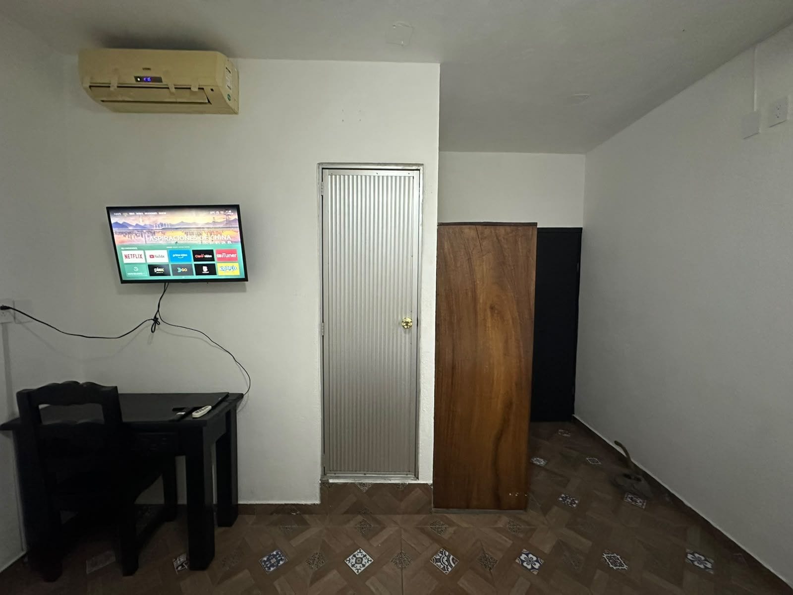 Una habitación con una televisión montada en la pared, una puerta, una mesa con una silla y suelo con diseño.