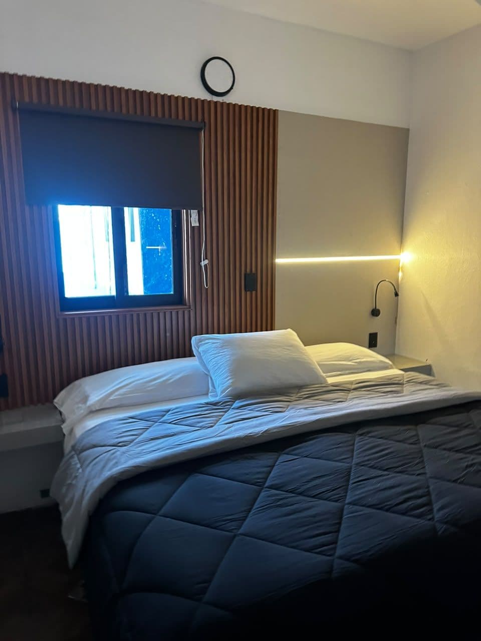A hotel room with a bed, a window with blinds, a bedside lamp, and a wall-mounted light.