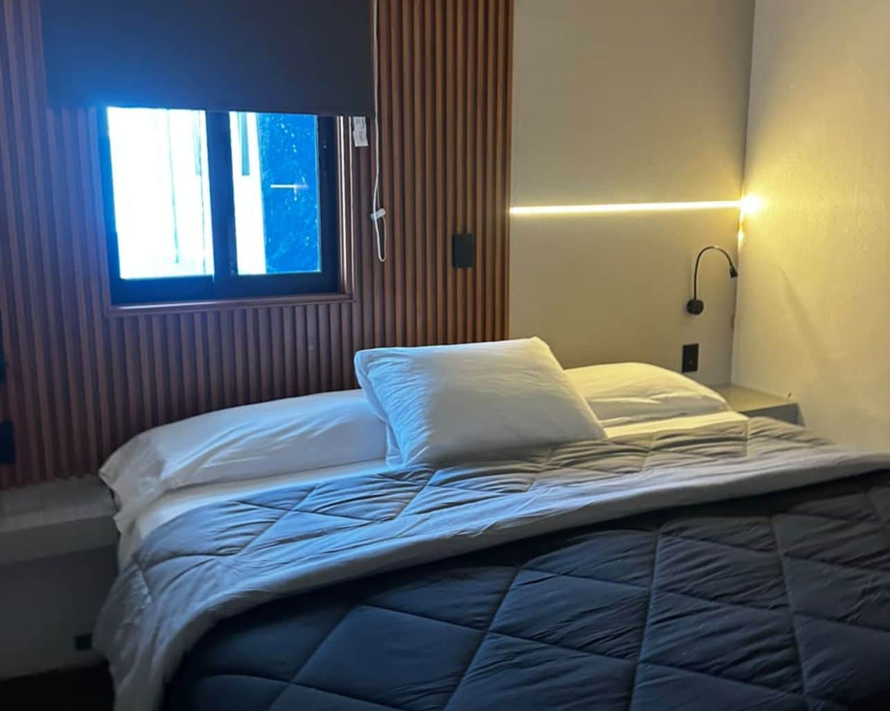 Una cama individual con cubrecama acolchado, una mesita de noche con lámpara y una ventana con persianas.