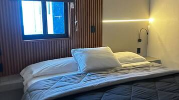 Una cama individual con cubrecama acolchado, una mesita de noche con lámpara y una ventana con persianas.