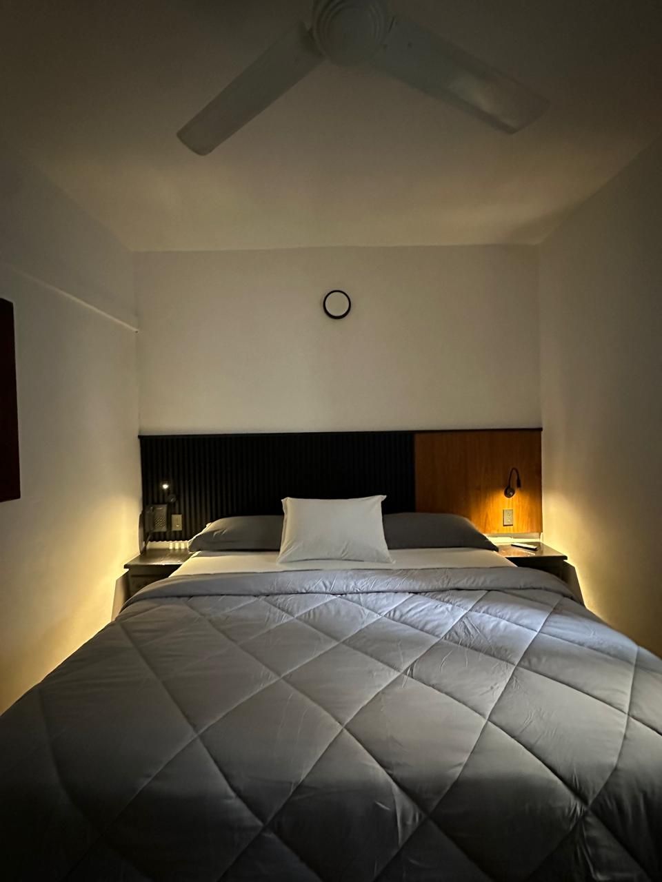 A hotel room with a bed, a nightstand, a wall clock, and a ceiling fan.