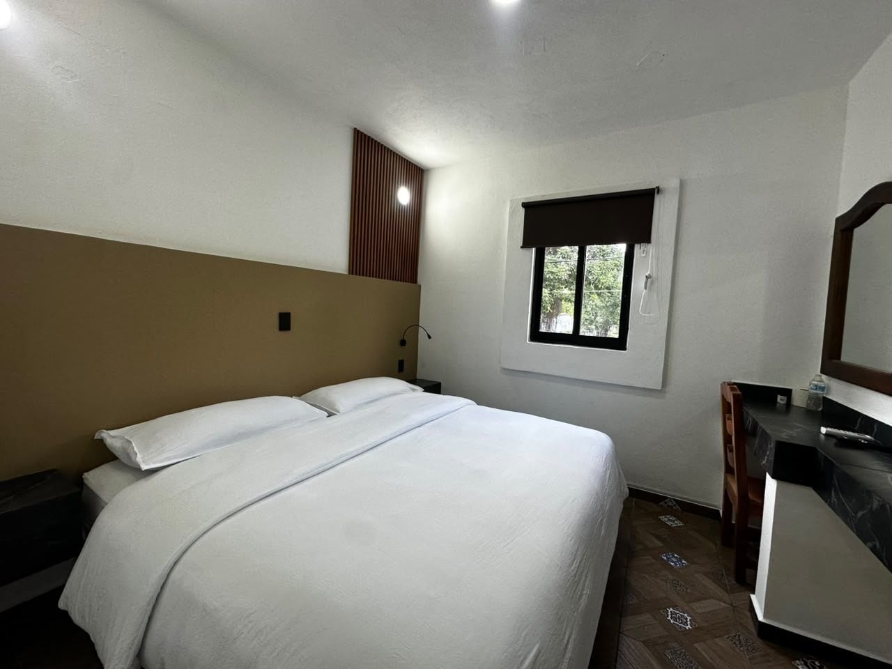 A bedroom with a bed, a nightstand, a desk, a mirror, and a window.
