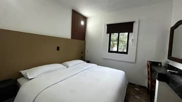 A bedroom with a bed, a nightstand, a desk, a mirror, and a window.