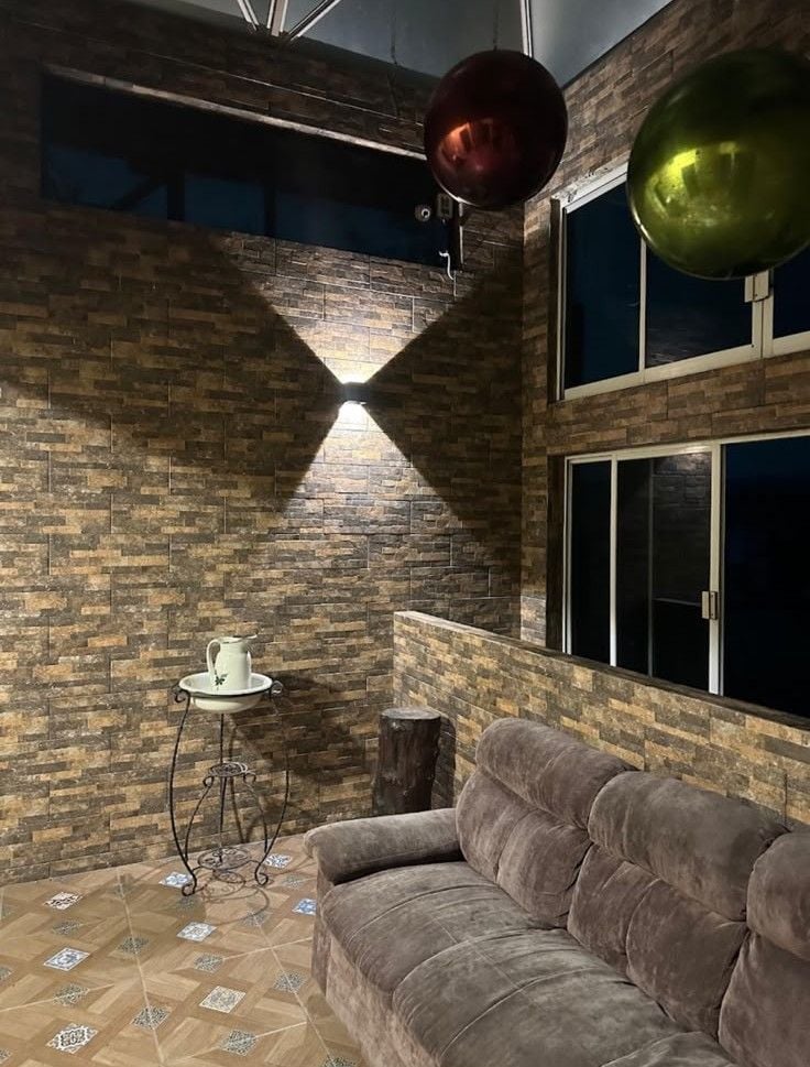 A room with a brick wall, a window, a sofa, a small table with a pitcher, and a hanging light fixture.