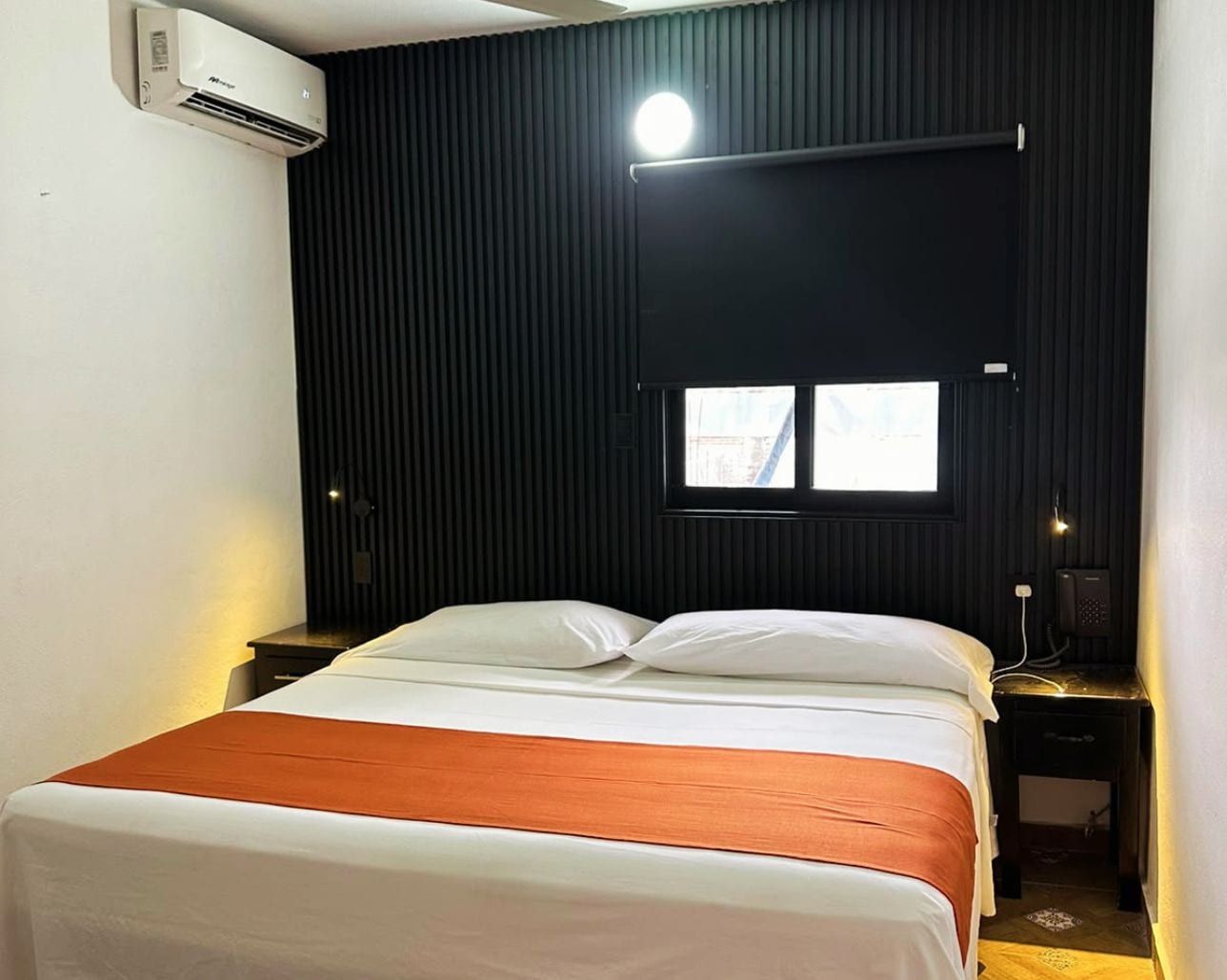 Habitación de hotel con cama, ventana, aire acondicionado de pared, mesita de noche con lámpara y teléfono.
