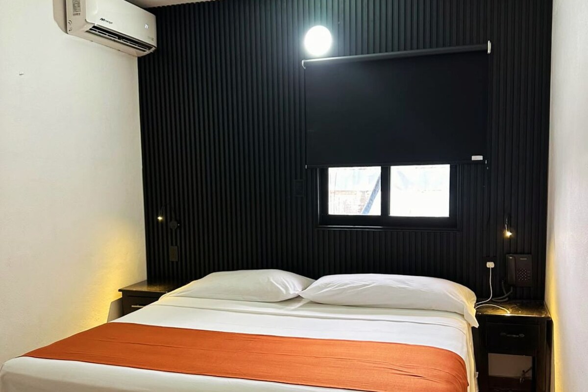 A hotel room with a bed, a window, a wall-mounted air conditioner, a bedside table with a lamp, and a telephone.