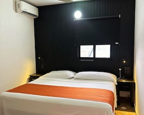 A hotel room with a bed, a window, a wall-mounted air conditioner, a bedside table with a lamp, and a telephone.