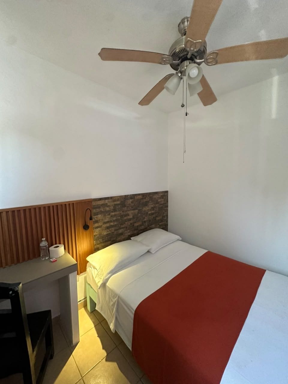 Una habitación con cama, escritorio, ventilador de techo e iluminación en la pared.