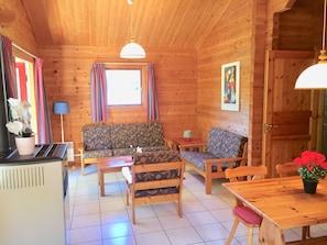 A wooden cabin interior with a sofa, chairs, a table, a lamp, and a window with curtains.