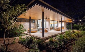 A modern house with a covered patio, stone walls, and a garden with various plants. - Seasons at Ceiba | Staffed Luxury House + Gym (Pinilla)