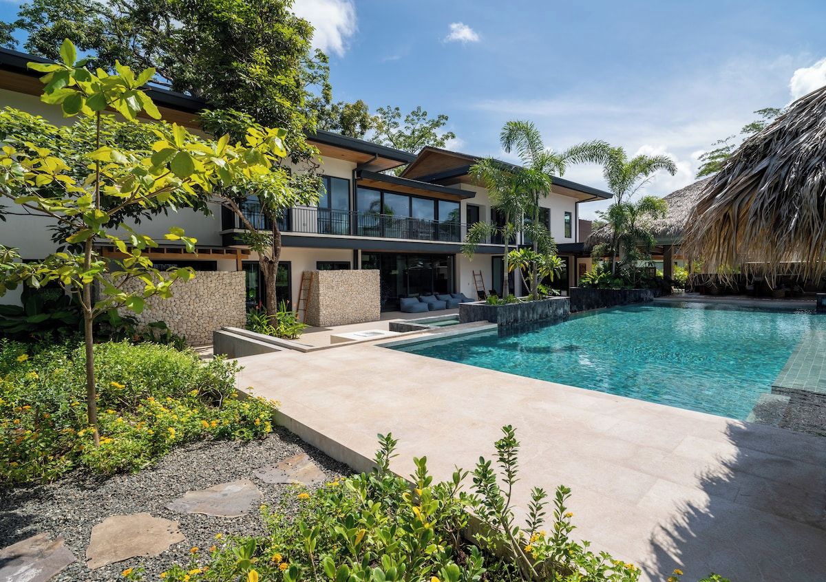 A modern house with a swimming pool, surrounded by tropical plants and a thatched roof structure.