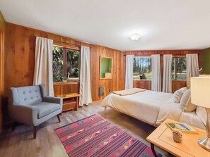 Ein Schlafzimmer mit einem Bett, einem Sessel, einem Nachttisch mit Lampe, einem Fenster mit Vorhängen und einem gemusterten Teppich.