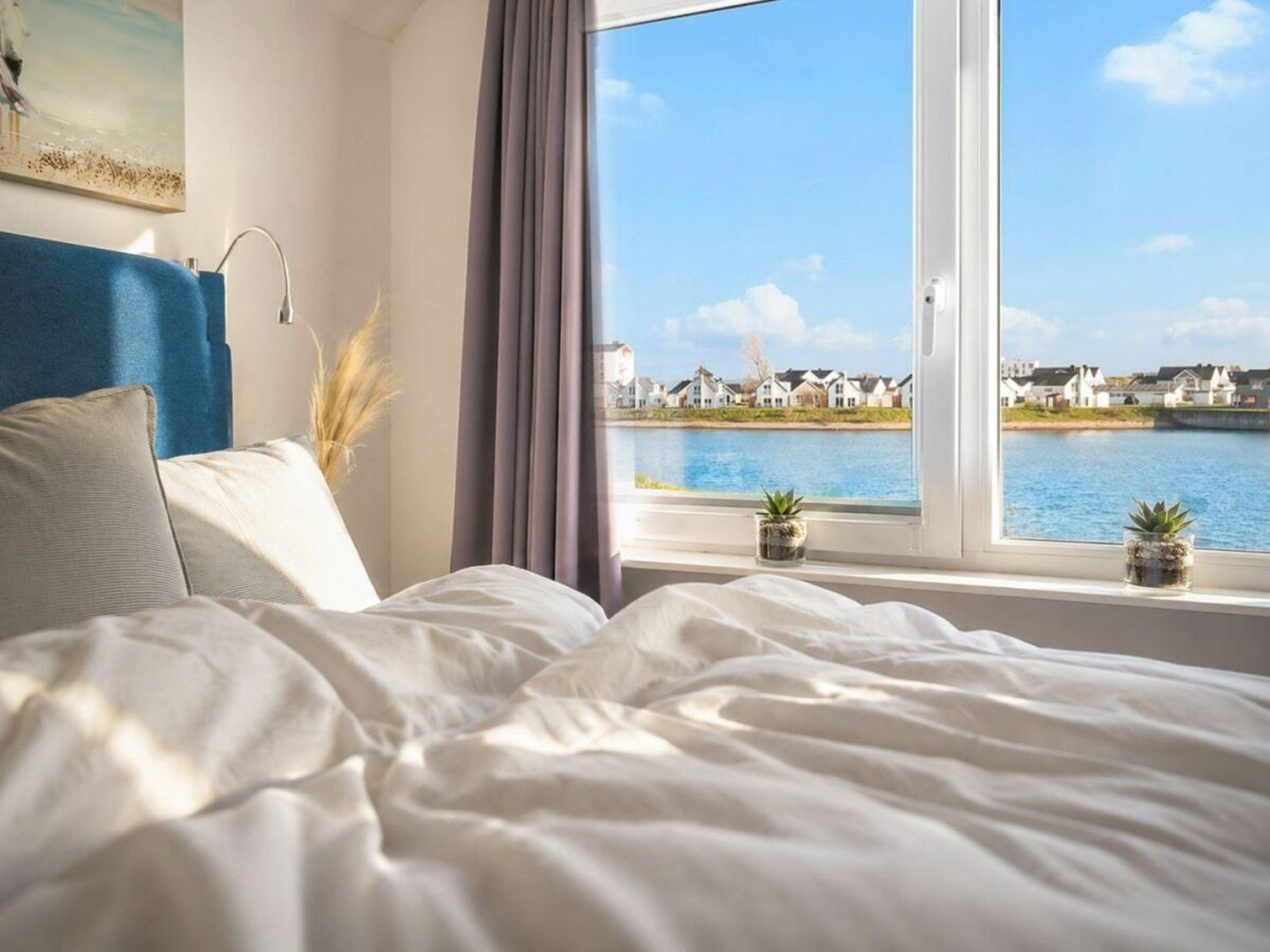 A bedroom with a large window, a bed with white bedding, a blue headboard, and two small potted plants on the windowsill.