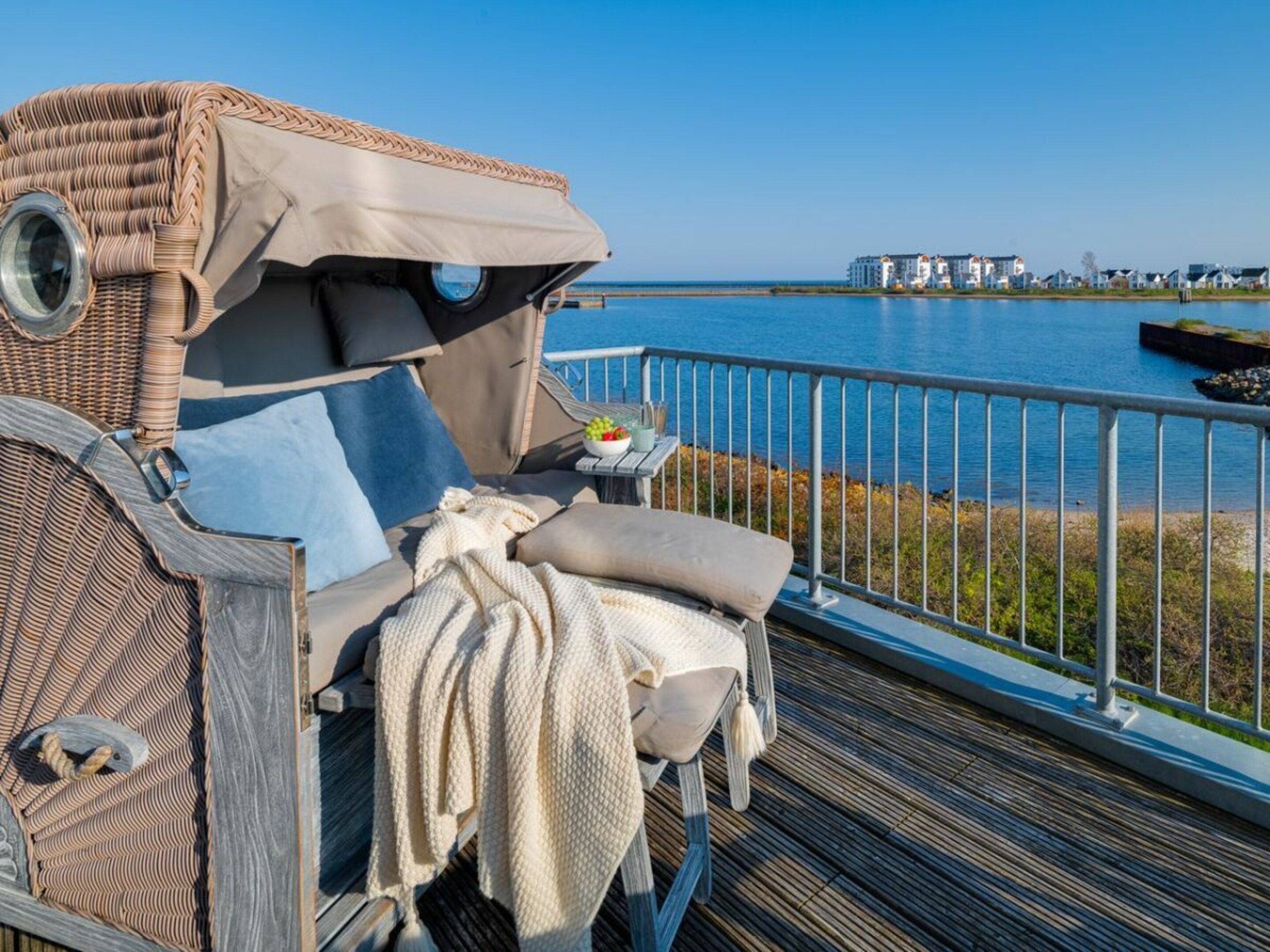 A deck with a lounge chair, a small table with a bowl of fruit, and a view of a body of water and buildings in the distance.