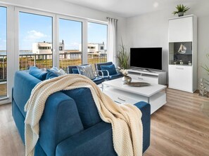 A modern living room with a sofa, coffee table, TV, and a view of buildings through large windows. - Holiday Apartment With 1 Bedroom (Kappeln)