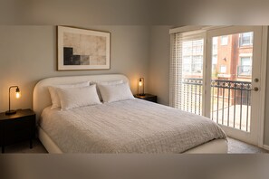 A bedroom with a bed, two bedside lamps, a framed artwork on the wall, and a sliding glass door leading to an outdoor balcony. - SUBLET — LUXURY FURNISHED RENTAL IN ENGLEWOOD, NJ (Englewood)