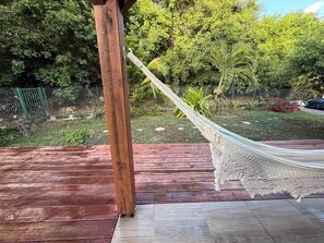 A hammock on a wooden deck with a view of a lush garden and a green fence. - To complete (Saint-Francois)