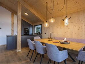 Ein moderner Essbereich mit Holztisch, grauen gepolsterten Stühlen, Pendelleuchten und Blick in die Küche durch ein Fenster.
