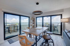 A modern interior with a wooden dining table, chairs, a sofa, and large windows offering a view of the sea. - Bay Views & Heated Pool | Coastal 2BR Retreat (Bradenton Beach)