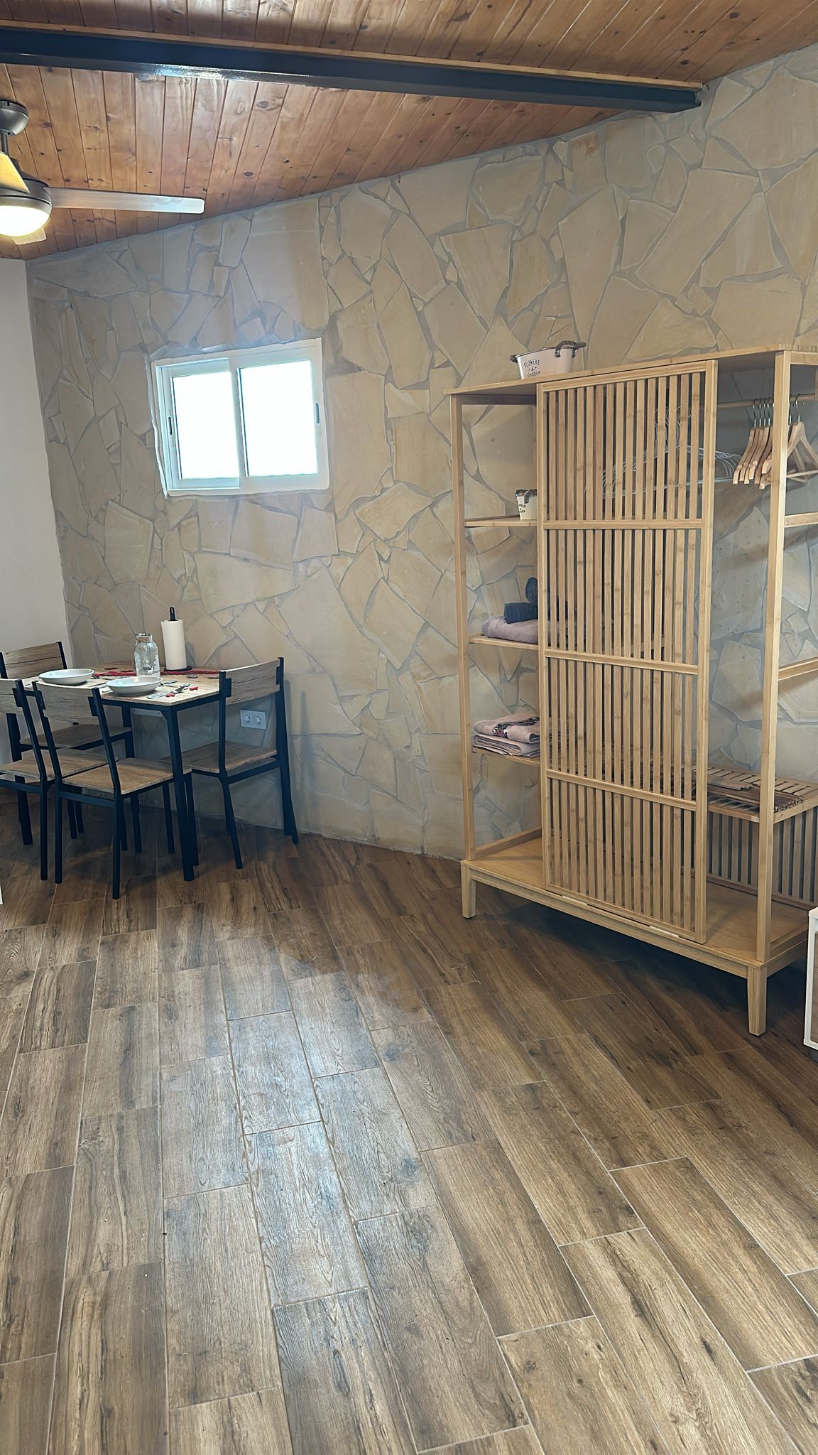 A room with a dining table, chairs, a wooden wardrobe, and a stone wall.