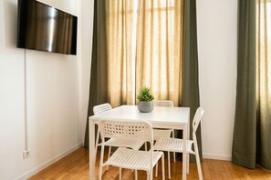Ein Zimmer mit einem weißen Tisch, vier weißen Stühlen, einer Zimmerpflanze, einem flachbildfernseher an der Wand und grünen Vorhängen.