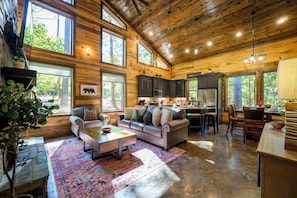A spacious living room with a large sofa, a coffee table, and a rug. There is a kitchen area with dark cabinets and a dining table. The room has large windows and a wooden ceiling. - 3 Private King Suites | Hot Tub, Playset, PingPong (Broken Bow)