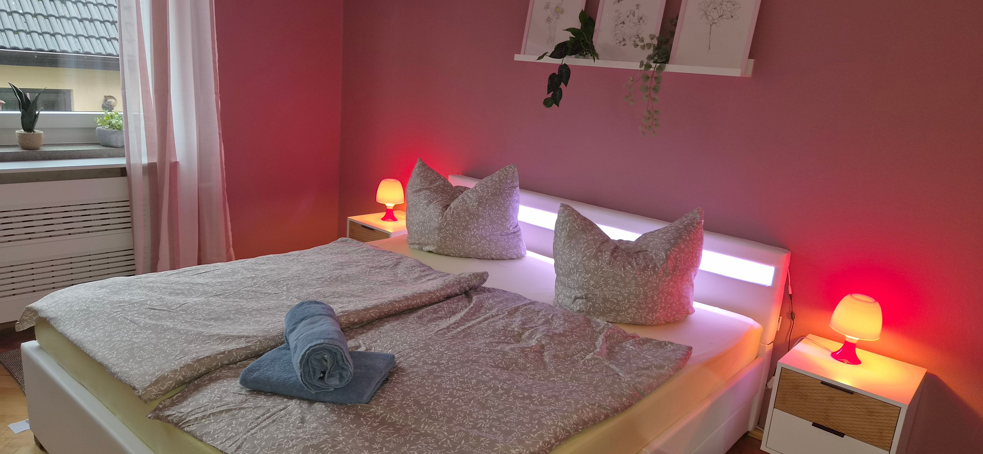 Ein ordentlich bezogenes Bett mit gemusterten Bettwäsche und Kissen, eine Nachttischlampe und ein Fenster mit Blick nach draußen.