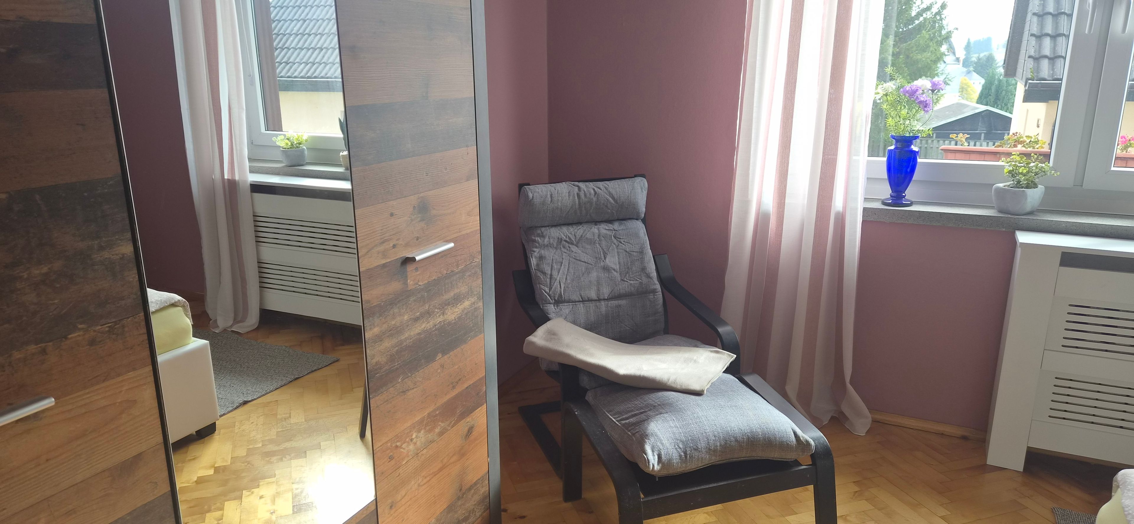 Una habitación con piso de madera, un sillón tapizado gris con cojín, una ventana con cortinas y una pequeña maceta en el alféizar.