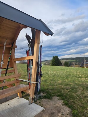 Een houten constructie met ski-uitrusting, een houten trap en uitzicht op een groen veld en heuvels in de verte.