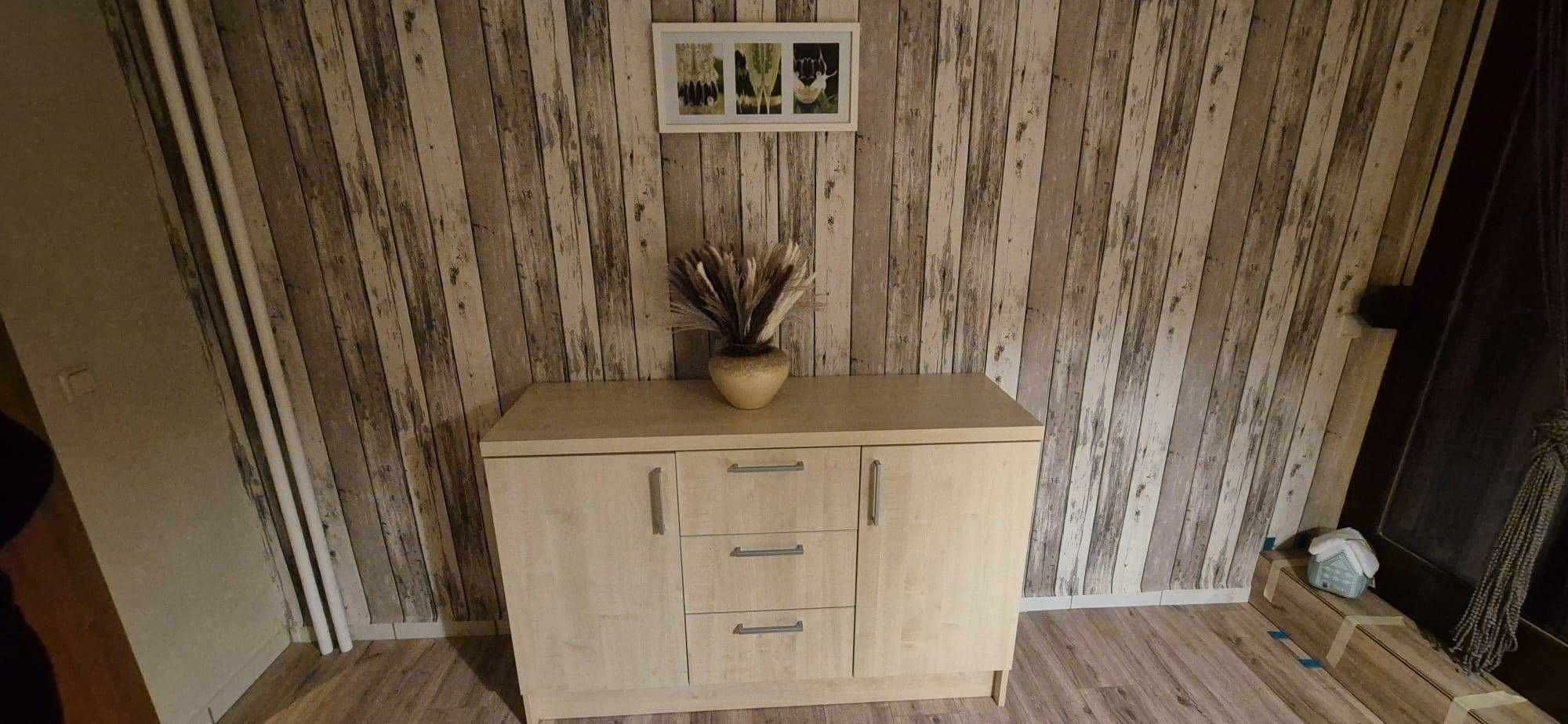 Un mueble de madera con tres cajones, un florero con plantas secas en la parte superior y un cuadro en la pared.