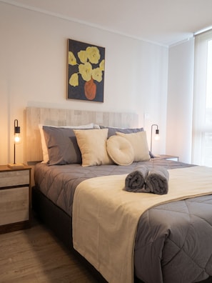 A neatly made bed with a beige and gray duvet, two bedside lamps, and a wall-mounted painting of flowers. - Edificio Arturo Prat by Domus Travels (Santiago)