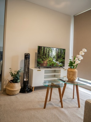Una sala de estar con un televisor de pantalla plana sobre un mueble blanco, una lámpara de pie alta, una mesa de centro de vidrio y dos cestas tejidas con plantas.