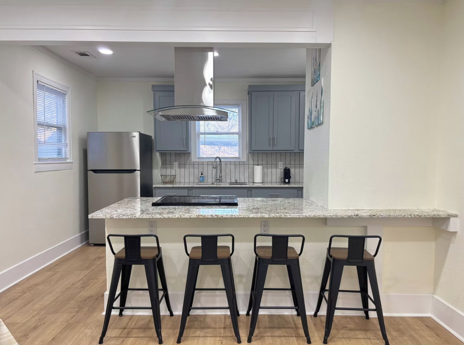 A modern kitchen with a central island, bar stools, a refrigerator, and a window with blinds.