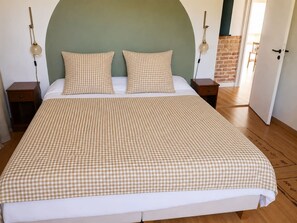 Una cama con colcha a cuadros, dos almohadas, mesitas de noche con lámparas y una pared de ladrillo al fondo.