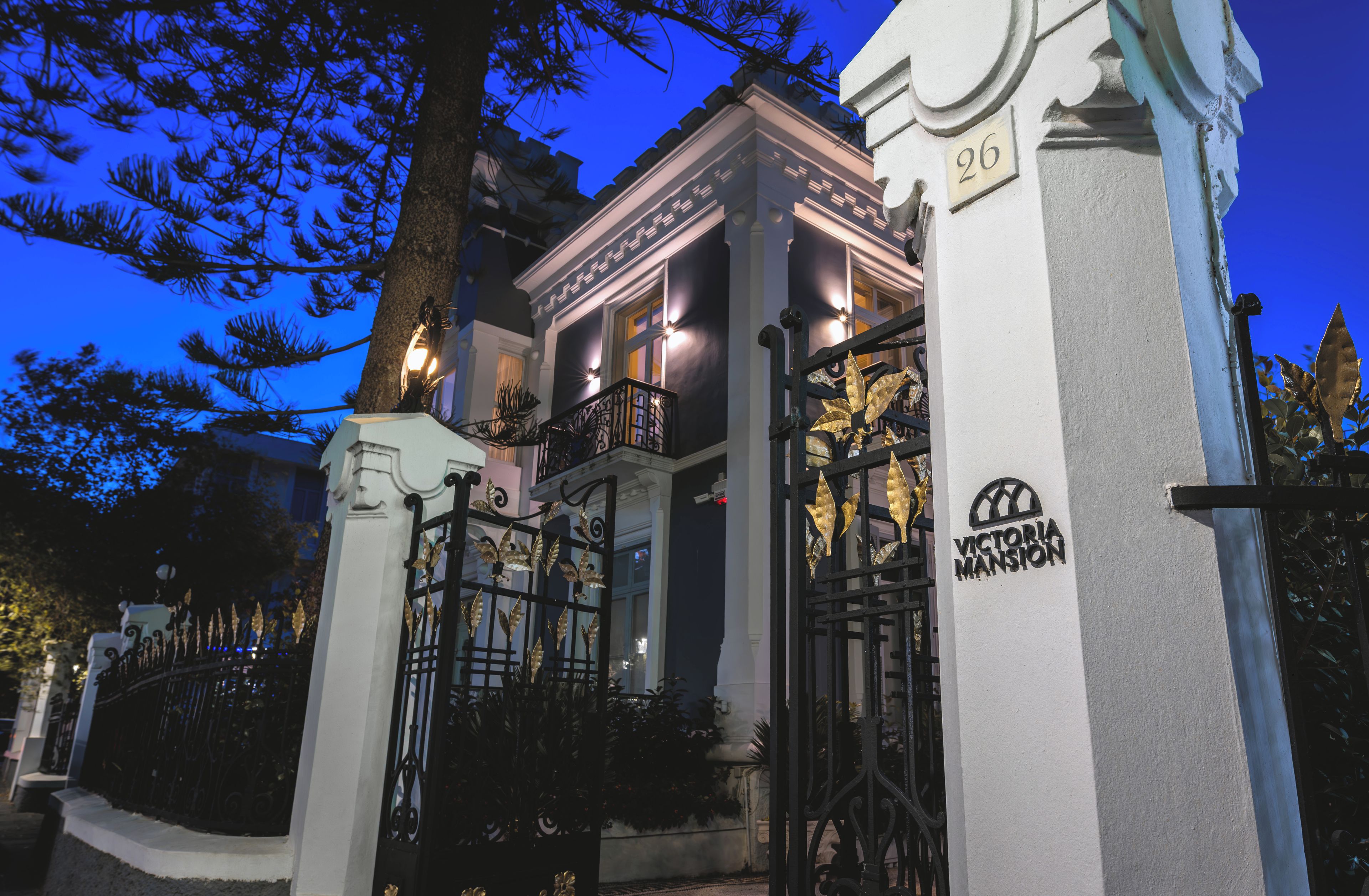 Un edificio con entrada protegida por una reja, un balcón con ornamentación metálica y un cartel que indica el nombre 'VICTORIA MANSION'.