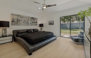 Ein modernes Schlafzimmer mit einem großen Bett, einem Deckenventilator und Blick auf einen Garten durch eine Schiebetür.